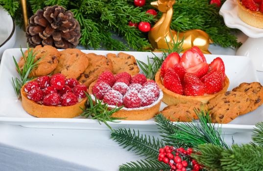 Plateau de desserts avec tartes aux fruits rouges, biscuits et décorations festives en arrière-plan.